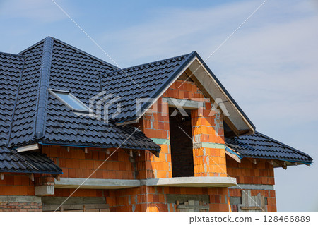 Metal tile roofing is being installed on a partially built house under a clear blue sky, showcasing modern construction techniques Metal tile roofing is being installed on a partially built house under a clear blue sky, showcasing modern construction techniques 128466889