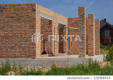 Brick walls are being constructed at a building site, with a pallet jack visible, surrounded by greenery under a clear blue sky Brick walls are being constructed at a building site, with a pallet jack visible, surrounded by greenery under a clear blue sky 128466918