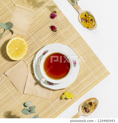 Tea time. Healing natural herbal tea. Sustainable morning composition with cup of herbal tea with spoons, slice of lemon and dry flowers on wooden napkin on white background. Flat lay 128466943
