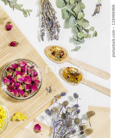Home herbal apothecary concept. Dry flowers and herbs, tea bags and wooden spoons on wooden napkin on white background. Healing tea from wild plants and flowers. Flat lay, vertical format 128466966