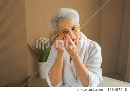 Eye patch beauty aging treatment. Mature adult old woman with under eye collagen pads. Senior woman with patches under eyes gets facial treatment. Senior lady in robe relaxing in bathroom at home Eye patch beauty aging treatment. Mature adult old woman with under eye collagen pads. Senior woman with patches under eyes gets facial treatment. Senior lady in robe relaxing in bathroom at home 128467048