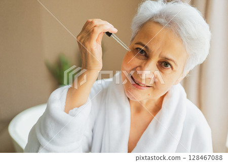 Woman applying natural drops cosmetics oil to face skin. Mature adult old woman applying nourishing anti age serum with pipette doing daily beauty routine. Senior woman gets kin scare facial treatment Woman applying natural drops cosmetics oil to face skin. Mature adult old woman applying nourishing anti age serum with pipette doing daily beauty routine. Senior woman gets kin scare facial treatment 128467088