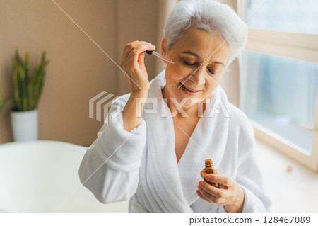 Woman applying natural drops cosmetics oil to face skin. Mature adult old woman applying nourishing anti age serum with pipette doing daily beauty routine. Senior woman gets kin scare facial treatment Woman applying natural drops cosmetics oil to face skin. Mature adult old woman applying nourishing anti age serum with pipette doing daily beauty routine. Senior woman gets kin scare facial treatment 128467089