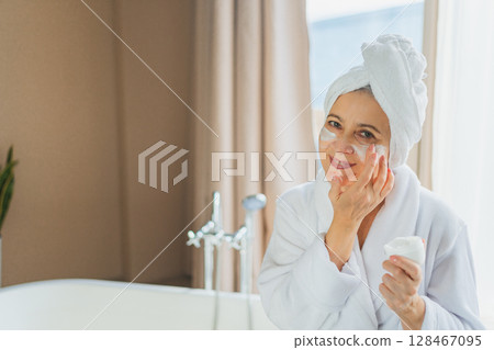 Mature adult old woman applying nourishing anti age face cream doing daily beauty routine. Senior woman with white mask or creme on skin gets facial treatment Skin care aging beauty skincare cosmetics 128467095