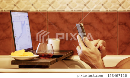 Man trading digital currencies online while relaxing in bathtub with laptop, ipad and a glass of cognac. Online work at home. Relaxing trader 128467116