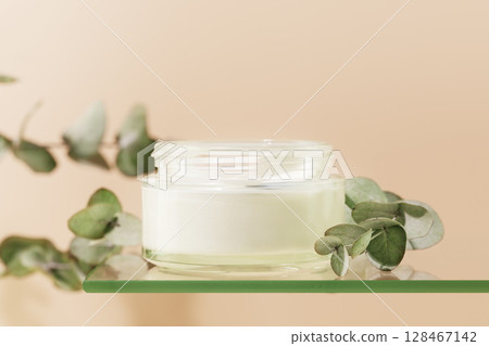 Cosmetic glass jar with natural white cream on glass shelf on pink background with eucalyptus sprigs. Organic beauty product and skin friendly cosmetics concept. Mockup, front view, close up 128467142