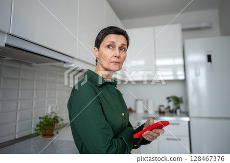 Upset lost mature woman holding smartphone in kitchen after receiving bad news. Emotional distress 128467376