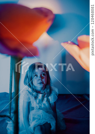 Child sitting among illuminated flowers in a softly lit indoor setting during evening 128468081