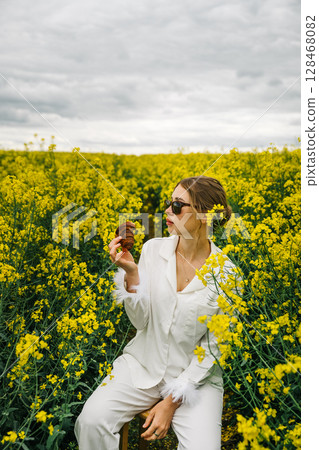Woman enjoying snack while sitting amidst vibrant yellow flowers in a field 128468082