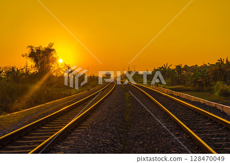 A golden sun dips towards the horizon, casting a warm glow over a curving railway track flanked by lush vegetation under a vibrant orange sky. 128470049