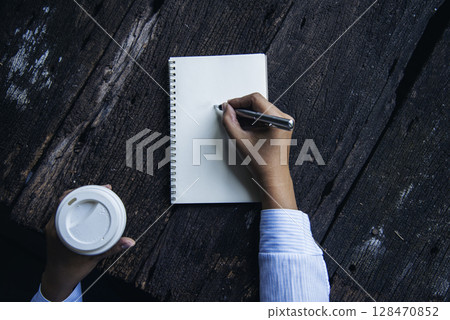 Woman hands writing empty notebook planing meeting agenda on wooden table. Close up women hands using pen drinking coffee cup planning office paper. Female hand holding pen note Personal Organizer Woman hands writing empty notebook planing meeting agenda on wooden table. Close up women hands using pen drinking coffee cup planning office paper. Female hand holding pen note Personal Organizer 128470852
