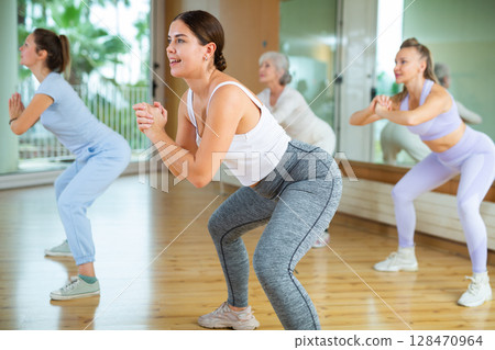 Group of positive sporty women of different ages in activewear performing squats before dance class with trainer in hall 128470964