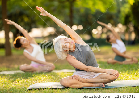 Sporty old lady sitting on sports mat and making yoga meditation in lotus pose and hands up with group of women together in green park at daytime 128471015