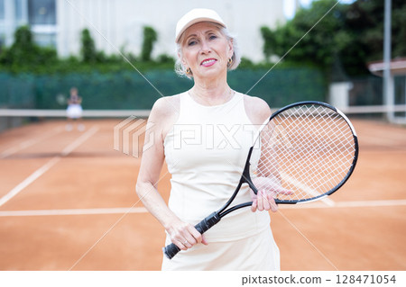 Senior woman in sportswear against backdrop of outdoor tennis court. Senior woman in sportswear against backdrop of outdoor tennis court. 128471054