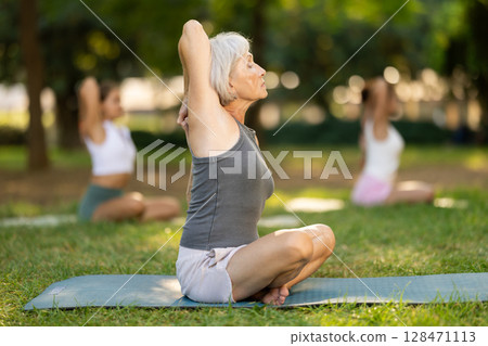 Sporty old lady sitting on sports mat and making yoga meditation in lotus pose and hands up with group of women together in green park at daytime Sporty old lady sitting on sports mat and making yoga meditation in lotus pose and hands up with group of women together in green park at daytime 128471113