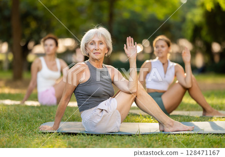 Senior woman performing Matsyendrasana during group yoga practice outdoors 128471167