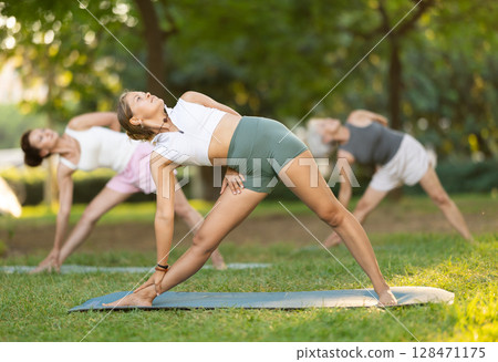 Sporty energetic females doing exercises on pilates mat during group training in park at daytime. Fitness concept 128471175