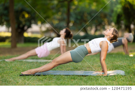 Girl doing Upward Plank Pose during group yoga in park 128471195