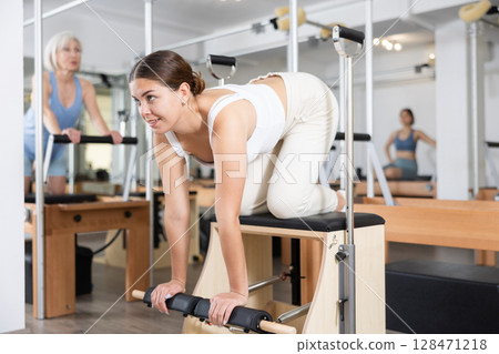 Young girl practicing pilates stretching exercises on combo chair Young girl practicing pilates stretching exercises on combo chair 128471218