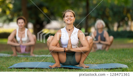 Active young girl teacher performs garland pose with group in green park Active young girl teacher performs garland pose with group in green park 128471361