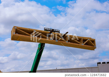 Crane hoists large wooden truss skyward against backdrop of works day indicating construction activity. 128471682