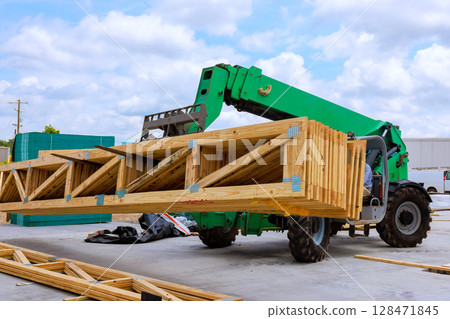 Construction vehicle is lifting large wooden truss beams at job site under during works daytime. 128471845