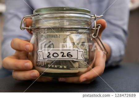 Unrecognizable woman holding Saving Money In Glass Jar filled with Dollars banknotes. 2026 year transcription in front of jar. Managing personal finances extra income for future insecurity 128472689