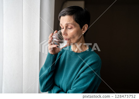Sad calm woman sip water at window lost in thought isolated indoors. Nervous tension, self soothing 128473752