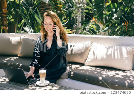 Happy Woman on Phone Call Working Remotely on Laptop Outdoors in Tropical Garden with Coffee Happy Woman on Phone Call Working Remotely on Laptop Outdoors in Tropical Garden with Coffee 128474630