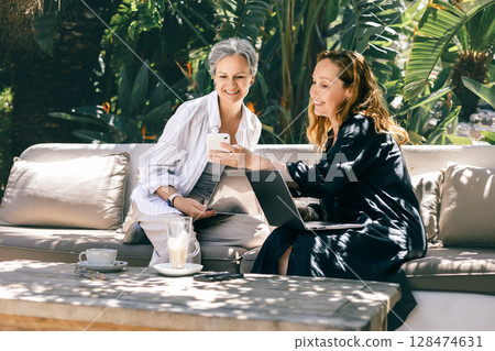 Two mature women working with laptops and smartphone outdoors, sharing ideas in a tropical setting Two mature women working with laptops and smartphone outdoors, sharing ideas in a tropical setting 128474631