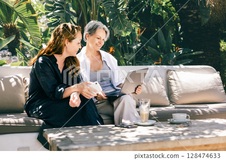 Happy mature women working remotely together with laptop and phone in tropical garden setting Happy mature women working remotely together with laptop and phone in tropical garden setting 128474633