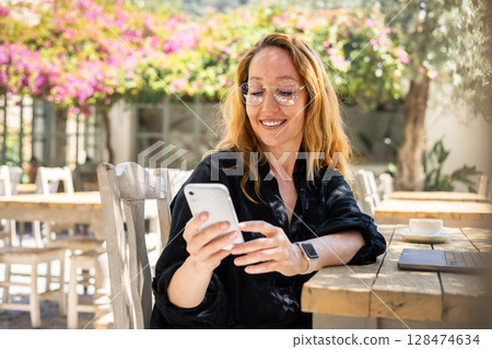 Smiling Woman Using Smartphone at Outdoor Cafe Table with Laptop and Coffee on a Sunny Day Smiling Woman Using Smartphone at Outdoor Cafe Table with Laptop and Coffee on a Sunny Day 128474634