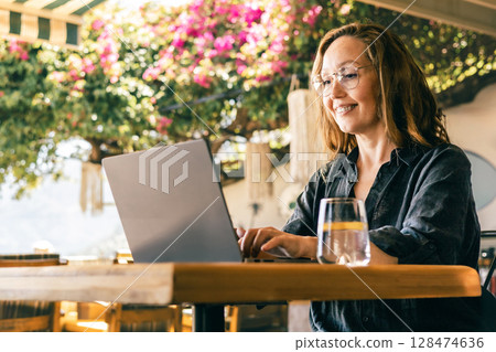 Happy Woman Working on Laptop Outdoors Under Blooming Tree in Sunny Cafe Environment 128474636