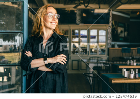 Confident Smiling Woman with Smartwatch Standing in Stylish Cafe Entrance 128474639