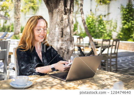 Smiling woman working remotely with laptop and smartphone in outdoor cafe on a sunny day 128474640