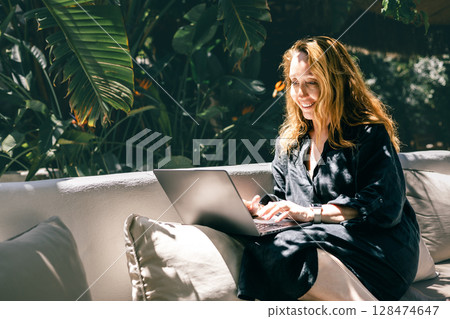 Happy Woman Working Remotely on Laptop in Tropical Outdoor Lounge with Sunlight and Greenery Happy Woman Working Remotely on Laptop in Tropical Outdoor Lounge with Sunlight and Greenery 128474647
