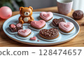 A delightful plate of heart shaped Valentines Day cookies and chocolates, decorated with pink and chocolate frosting, alongside a cute teddy bear figure, creating a charming and festive display. 128474657