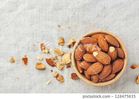 Roasted almonds in wood bowl with broken almond on linen tablecloth background, top view Roasted almonds in wood bowl with broken almond on linen tablecloth background, top view 128475635