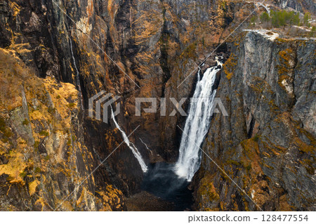 Magnificent Voringsfossen waterfall plunges into deep rugged canyon flanked by rocky cliffs. Winding road and sparse vegetation complete landscape 128477554