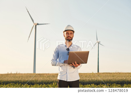 With laptop in hands. Service engineer is on the field with windmills With laptop in hands. Service engineer is on the field with windmills 128478019