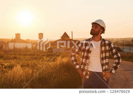 Man in hard hat is outdoors against sunset light. Rural scene 128478029