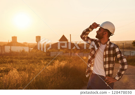 Touching the hard hat. Man is outdoors against sunset light. Rural scene Touching the hard hat. Man is outdoors against sunset light. Rural scene 128478030
