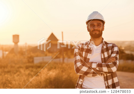 Man in hard hat is outdoors against sunset light. Rural scene Man in hard hat is outdoors against sunset light. Rural scene 128478033