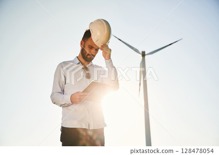 Standing against sunlight. Notepad in hands. Service engineer is on the field with windmills 128478054