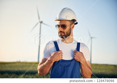 In blue colored uniform and in sunglasses. Service engineer is on the field with windmills In blue colored uniform and in sunglasses. Service engineer is on the field with windmills 128478098