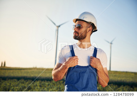 In blue colored uniform and in sunglasses. Service engineer is on the field with windmills 128478099