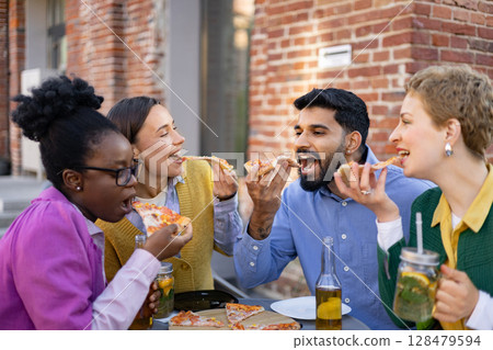 Group of friends joyfully eating pizza and drinks outdoors, enjoying a shared meal together at a restaurant or outdoor setting. 128479594