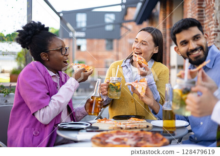 A group of friends enjoys pizza and drinks outdoors, laughing and sharing a meal together. A group of friends enjoys pizza and drinks outdoors, laughing and sharing a meal together. 128479619