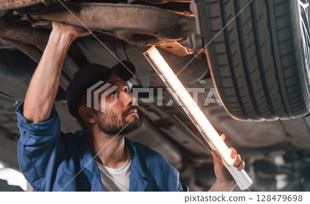 Under car, using lighting device. Auto mechanic working in garage. Repair service 128479698