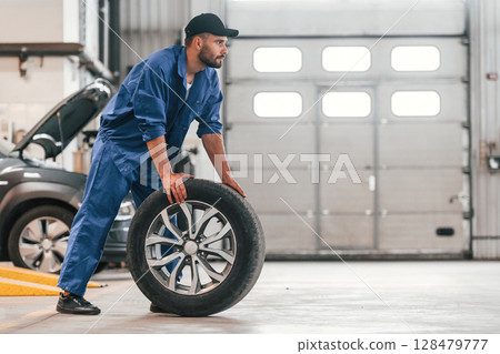 Side view. Moving the new tire. Auto mechanic working in garage. Repair service Side view. Moving the new tire. Auto mechanic working in garage. Repair service 128479777
